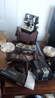 Group photo showing all four items: Brownie Automatic camera, Polaroid Land Camera with Q-Light flash, Keystone XR308 camera, Kodak Light Bar with dual reflectors