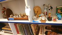 Wide view of wooden shelf holding Red Rose Tea Wade ceramic animal figurines arrayed in rows, bisque porcelain figurines including two human figures, miniature tea set with small cups and plates, and additional ceramic holiday figurines and decorations.