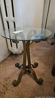 Full view of round glass top side table with metal base on carpeted floor, showing shape, size, and condition. Glass top is not attached to base.