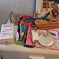 Wide shot showing multiple items on a dresser including manicure kits, glasses, a mirror, eye masks, a plastic tray, and wicker baskets holding assorted items.