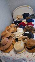 Photo of a bed covered with multiple ladies hats in various materials and colors, including wicker, straw, cotton, and felt, showing the quantity and variety of the collection.