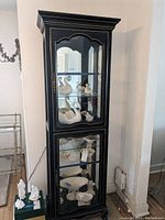 Tall black curio cabinet with four glass shelves visible through glass door and sides. Cabinet is lit internally with cord visible. The cabinet is placed in a corner against light-colored walls with some collectibles placed inside for display only.