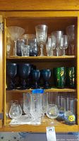 Wooden cabinet shelf with clear crystal stemware, dark blue goblets, short clear glasses, and green decorative glasses.