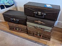 Six vintage 8 track storage cases stacked showing rectangular shape, different sizes, brown leather and fabric materials, metal clasps and handles.