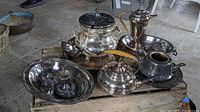 Overview of the assorted silver serving set including coffee urn, kettle, teapot, trays, and candle holders arranged on a wooden crate.