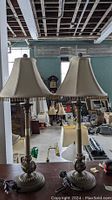 Pair of classic style table lamps with beige trapezoid fabric shades and fringe trim; metal column bases with decorative detailing.