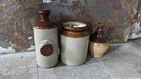 Set of three assorted ceramic jugs placed on a tiled floor against a textured wall, showing their shapes and colors.