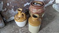 Three small brown and beige ceramic jugs positioned on a concrete surface with rustic wall background, showing distinct shapes and glazing.
