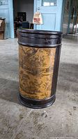 Front view of the cylindrical umbrella stand showing dark wood frame and yellow panels with scenic countryside illustration.