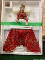 Porcelain holiday Barbie doll dressed in a red satin embroidered gown, placed in original white styrofoam packaging secured with green tape. Accessories include earrings and necklace, individually wrapped in foam.