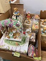 Wide view showing multiple Cherished Teddies figurines in original boxes, displayed in a cardboard box. Visible a large figurine depicting a bear on a swing under a pergola with purple flowers, along with several other boxed figurines.