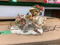 Front view of a Cherished Teddies Astrid figurine depicting a teddy bear in bonnet and dress standing next to a sleigh with smaller teddy bears and presents, with animal figures on snowy base