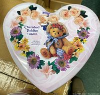 Close-up of a heart-shaped promo tin with floral design and the Cherished Teddies Cecilia teddy bear illustration on top.