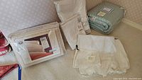 Photo showing folded bedding items on carpet including sealed packages and a lace dust ruffle.