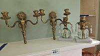 Two brass candle sconces with floral and leaf decorations, each with two candle holders, displayed hanging on a wall above a white mantle.