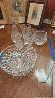 Six pieces of clear crystal and glass on wooden floor, includes decanter, large bowl, and four vases with a diamond cut pattern.