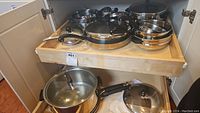 Full view of cookware in cabinet including frying pans, saucepans, and covered pots with handles.