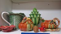 View of full lot showing the Spode Harvest creamer, sugar bowl with lid, salt and pepper shakers, green artichoke tulip vase, cabbage leaf pitcher, small red bird figurine, and pinecone figurine