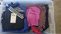 Top view of mixed folded women's tops including knit sweaters in various colors such as black, blue, pink, and striped patterns inside a plastic bin.