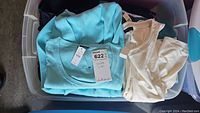 Plastic bin containing folded women's tops in size L-XL, showing light blue and cream tops with tags.