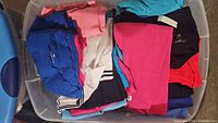 Clear photo showing a bin full of folded women's tops in various colors including blue, pink, white, black and red.