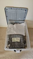 Open gray plastic bin showing folded women's pants including a pair of white pants and gray plaid pants with tags visible
