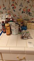 Photo showing various wood care liquids, repair markers, filler sticks, and Howard Restor-A-Finish cans against a floral wallpaper background on a tiled counter.