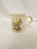 Front view of the small cream-colored ceramic pitcher with daisy and wheat decoration showing the rounded body, handle, and spout.