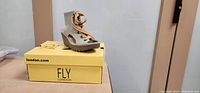 Side view of single beige open toed Fly London boot on top of yellow Fly London branded box.