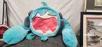 Large blue plush backpack shaped like Hatsune Miku's head laying on a couch, showing detailed hair, headphones and pink inner mouth area.