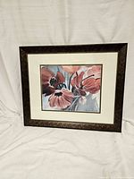 Photo of red floral framed print standing against a white fabric background, showing subtle decorative patterned dark frame and muted color matting.