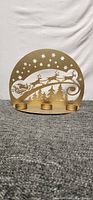 Front view of gold-tone metal tea light candle holder with cut-out winter scene including reindeer pulling Santa's sleigh, stars, and pine trees. The base holds three round platforms for tea lights.