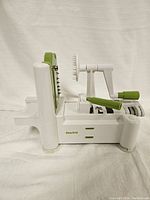 White and green Starfrit spiralizer with crank handle and blade attachments on a white cloth background.