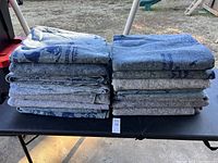 Two stacks of 14 folded gray moving blankets with blue printed text, placed on a black table outdoors on grass.