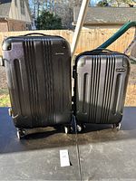 Photo of two black hard shell Rockland suitcases side by side on a surface outdoors with grass and fence in background.