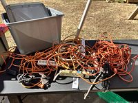 Large tangled pile of mainly orange extension cords with some power strips on a table, plastic storage bin in the background.
