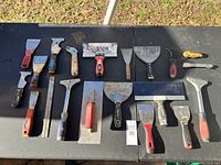 Photo showing 16 assorted hand tools arranged on a black surface including scrapers and putty knives of different sizes and handle types.