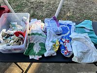Wide view of the lot spread out on a table, showing multiple folded and unfolded vintage table covers in various colors and patterns, some in a plastic bin.