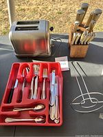 Photo showing stainless steel toaster, red tray with flatware, metal toast rack, and wooden knife block with knives.