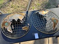 Two metal globe wall art pieces resting on a table outside. Both show continents in a grid wireframe sphere with rustic patinated metal plating.