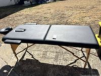 Wide angled side view of black padded massage table with wooden folding legs outdoors on concrete ground.
