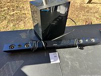 Samsung soundbar and subwoofer shown on table outdoors with power cable visible, lot card for identification