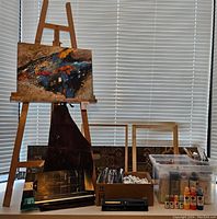 Full view of wood artist easel holding an abstract textured painting, surrounded by boxes and container of painting supplies and tools.