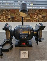 Front view of the bench grinder showing grinding wheels, safety guards, motor label, and cord.
