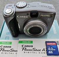 Front view of Canon PowerShot A710 IS camera showing lens, flash, mode dial, speaker grill, and branding with 4GB SanDisk SD card and manuals on box.