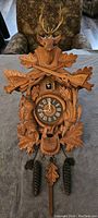 Front view of wood-carved cuckoo clock showing stag head, antlers, rifles, oak leaves, roman numeral dial, pinecone weights and leaf pendulum.