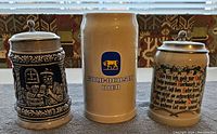Front view of three German beer steins showing logos, text and detailed relief with pewter lids