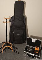 Full view of all items in lot including guitar soft case, two guitar stands, and aluminum case containing foot switch and effects pedal.