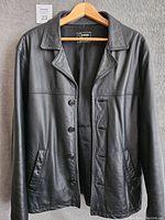 Front view of black leather car coat jacket on hanger showing button closure and two side pockets