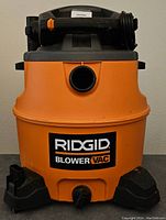 Front view of orange and black Rigid shop vacuum showing large tank and front intake port, power cord on top.